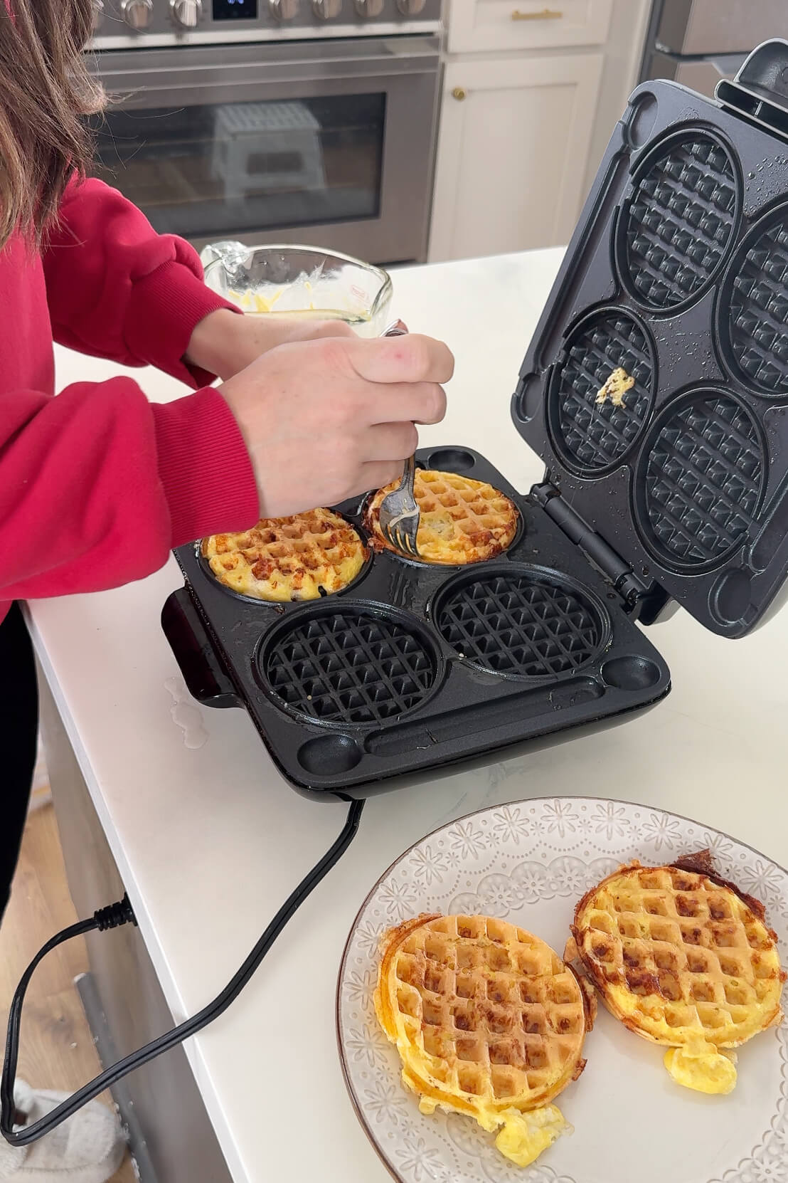 A woman in a red sweater uses a fork to lift a freshly cooked chaffle from the waffle maker, revealing its crispy, golden texture. Two finished chaffles rest on a plate beside her. Tha makings of a carnivore diet breakfast.